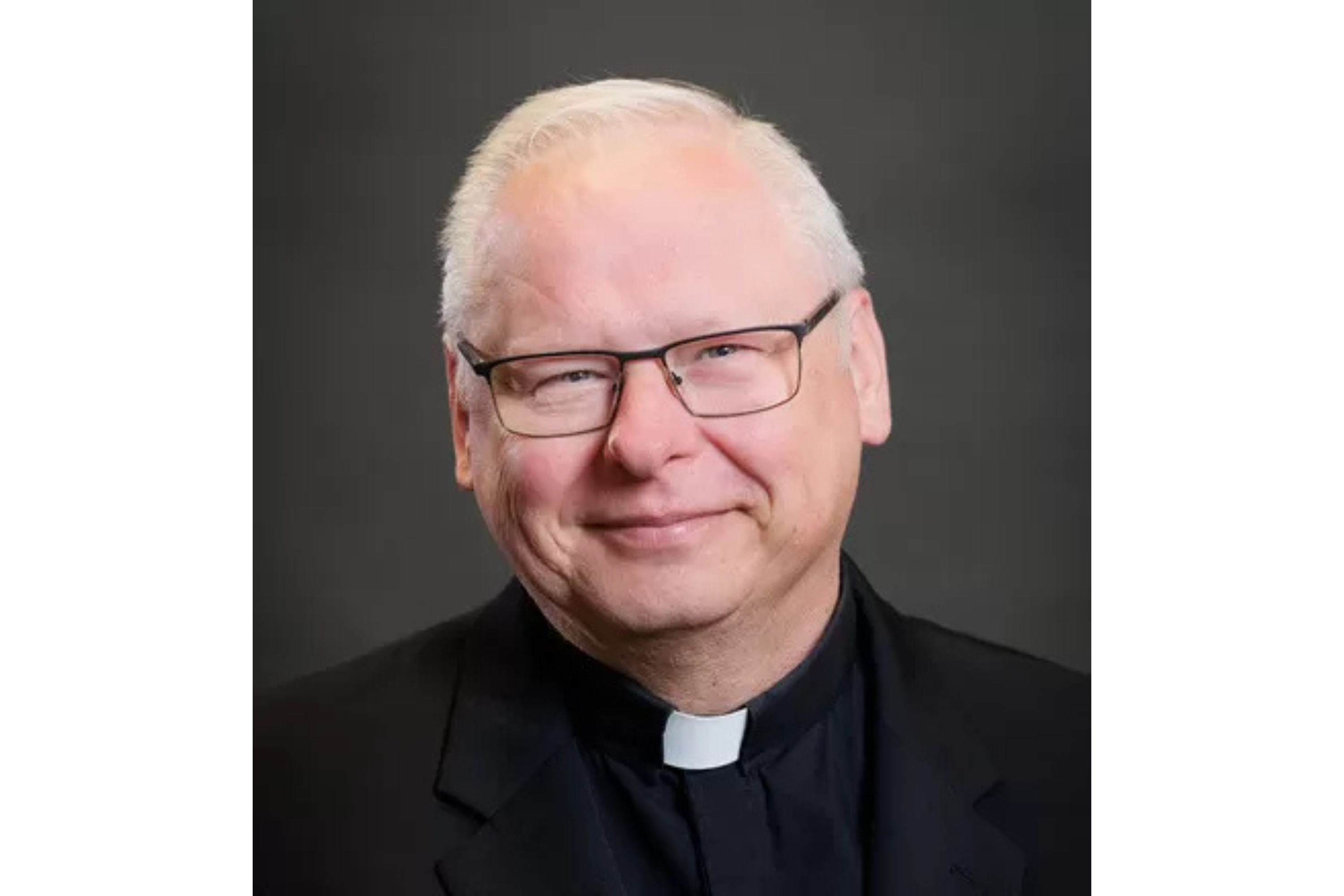 A white man with glasses and white hair smiles for a professional headshot. He wears the black shirt and white collar of a Catholic priest.