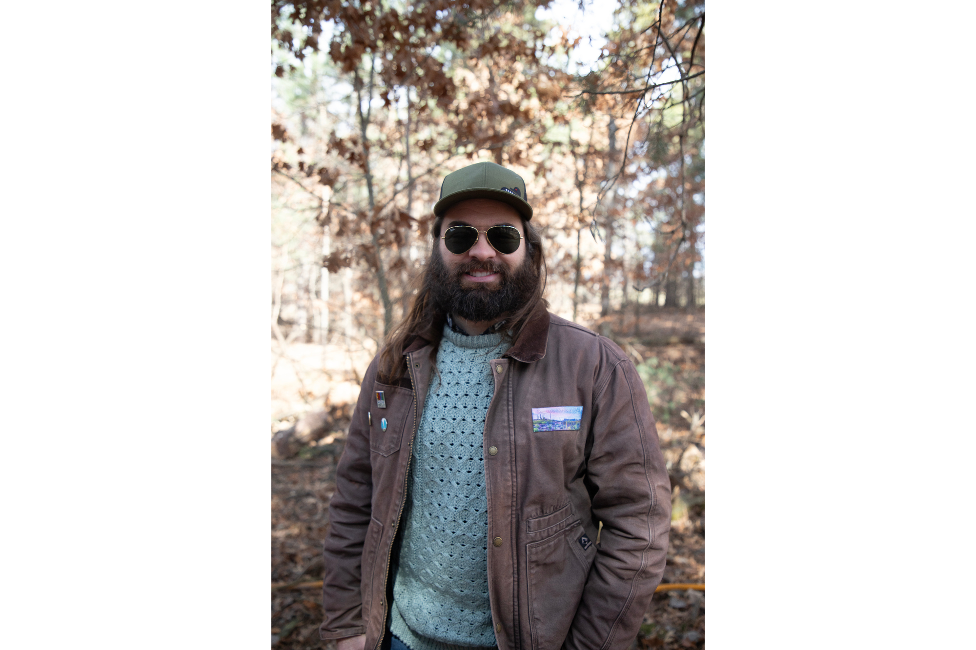 A man smiles for a portrait. He wears a dark green baseball cap, aviator sunglasses, and a brown coat over a green sweatshirt. He has long, dark hair and a dark beard. 