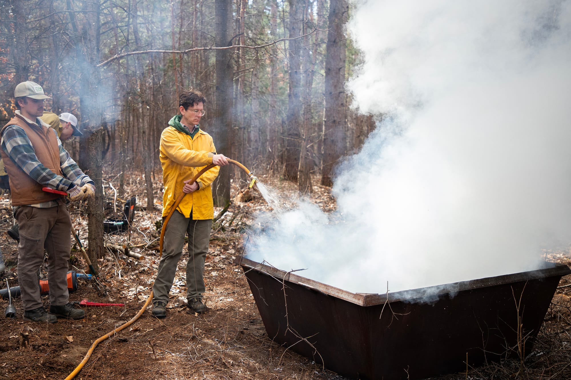 A man wearing a yellow coat uses a hose to quench a biochar kiln. The metal kiln sits on a forest floor, and smoke billows from it.