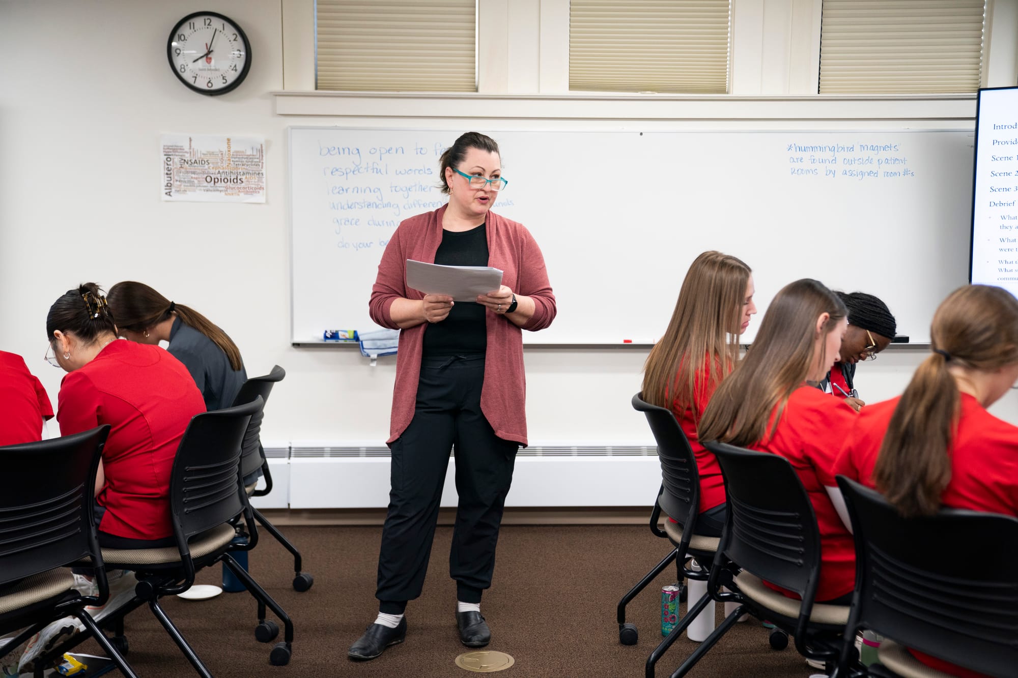 A woman wearing black pants, a black shirt, and a red sweater stands at the front of a class and reads from a sheet of paper. Students are seen from behind sitting in chairs looking at their desks. 