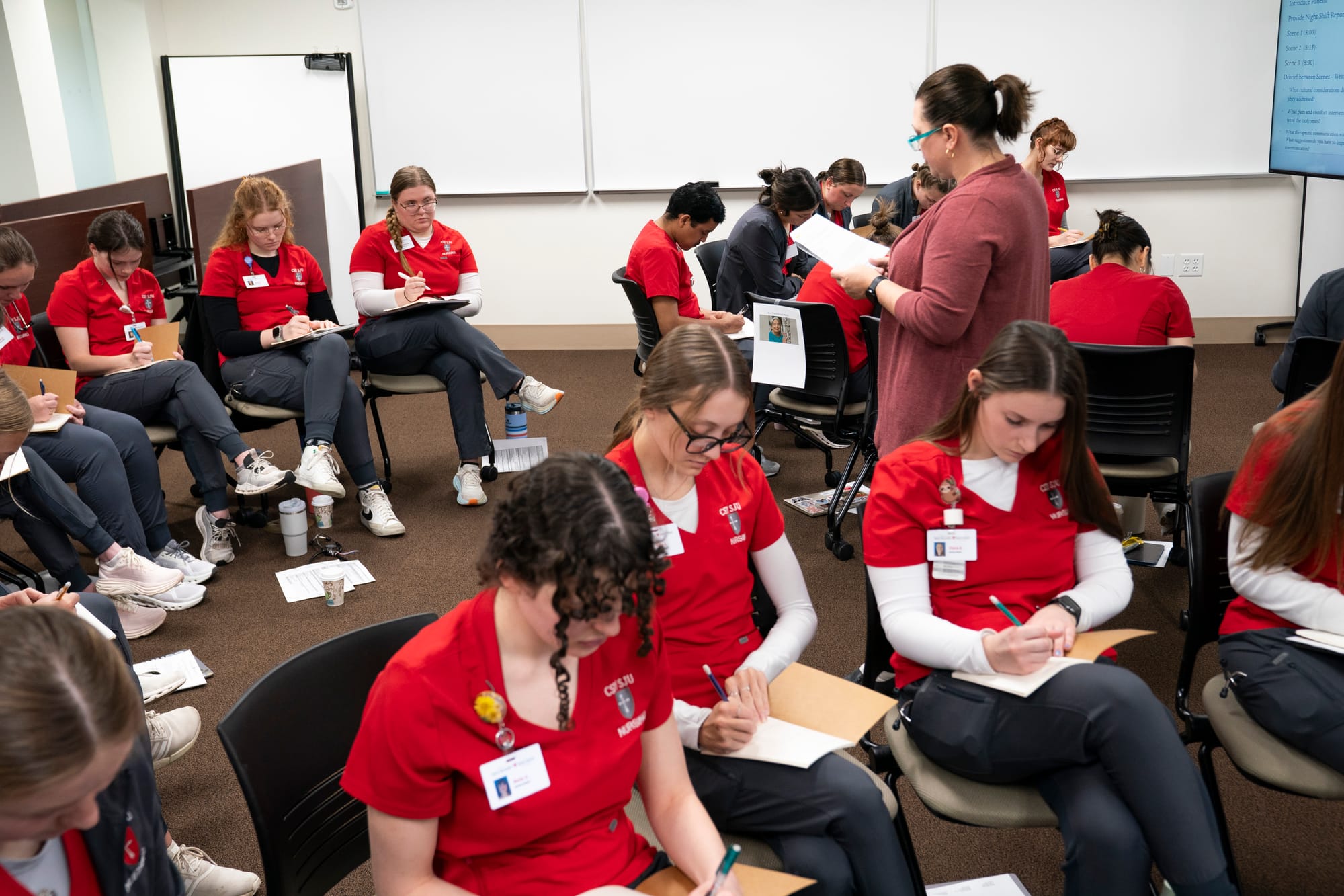 Nursing students are seen sitting on chairs and writing information on sheets of paper in front of them. An instructor stands in the upper left corner of the frame reading to them. 