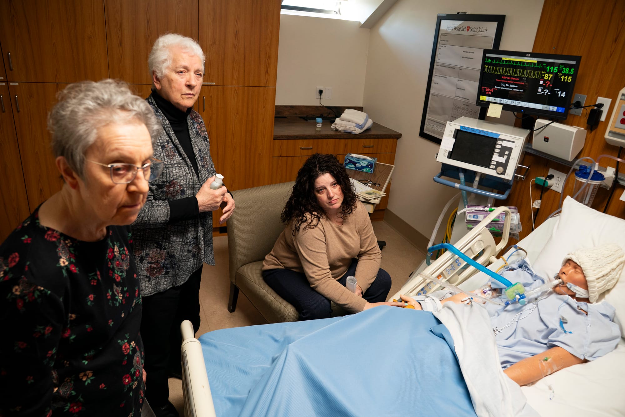 Three women stand and sit around a hospital bed. A mannequin used to train nursing students lays in the bed. 