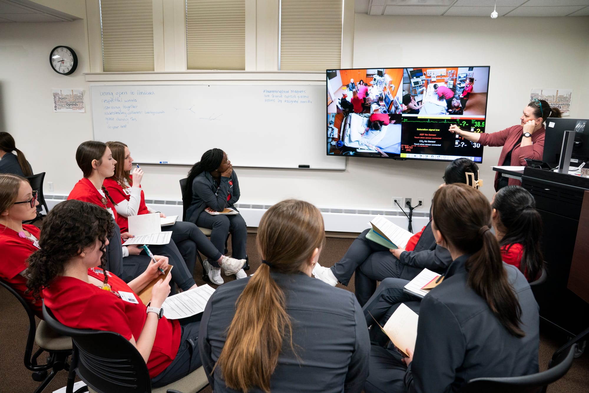 Nursing students are seen from behind watching a TV that shows their peers during an end-of-life training exercise. 