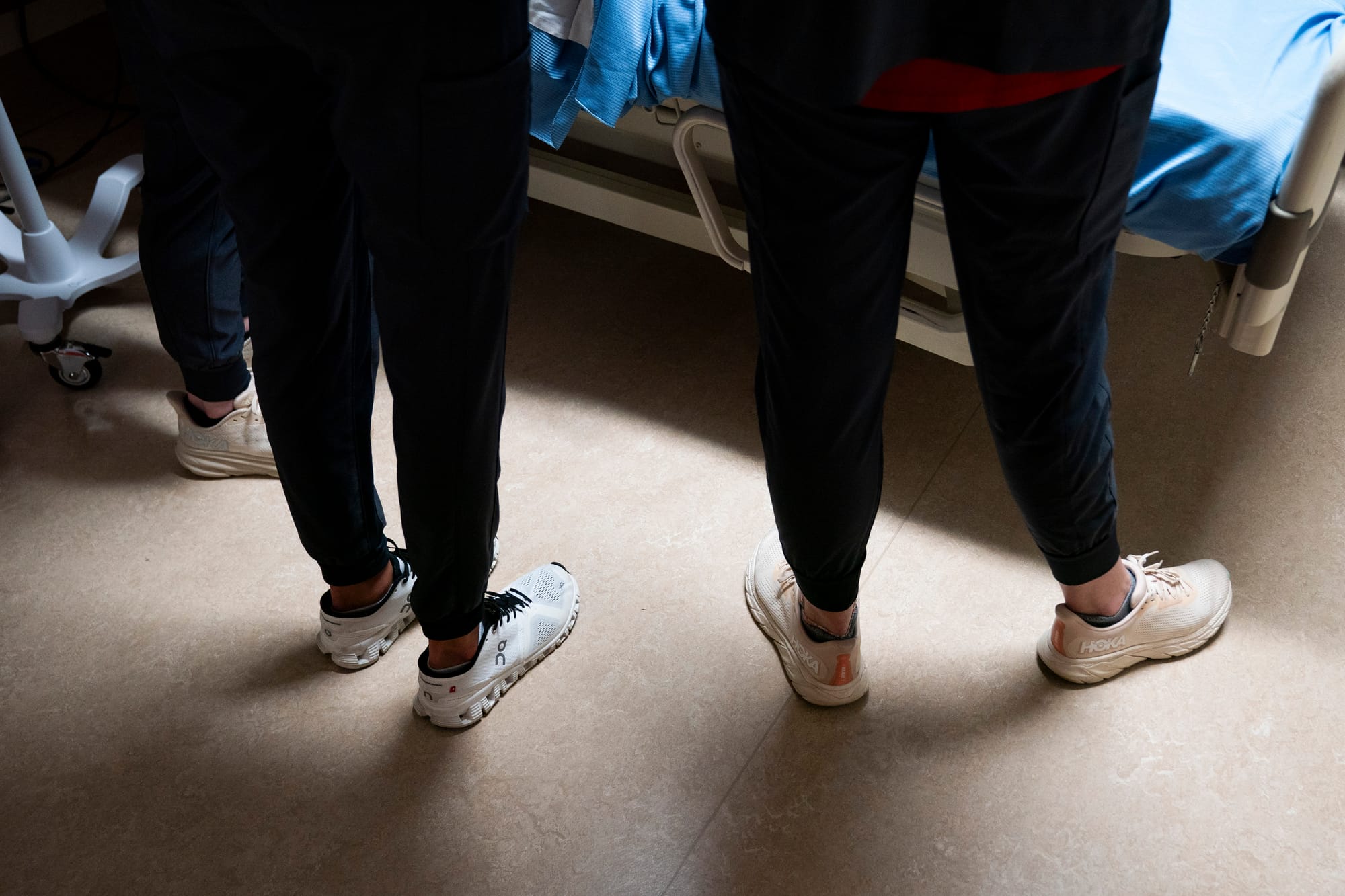 Nursing students stand next to a patient's bed during a training exercise. The photo shows their sneakers. 