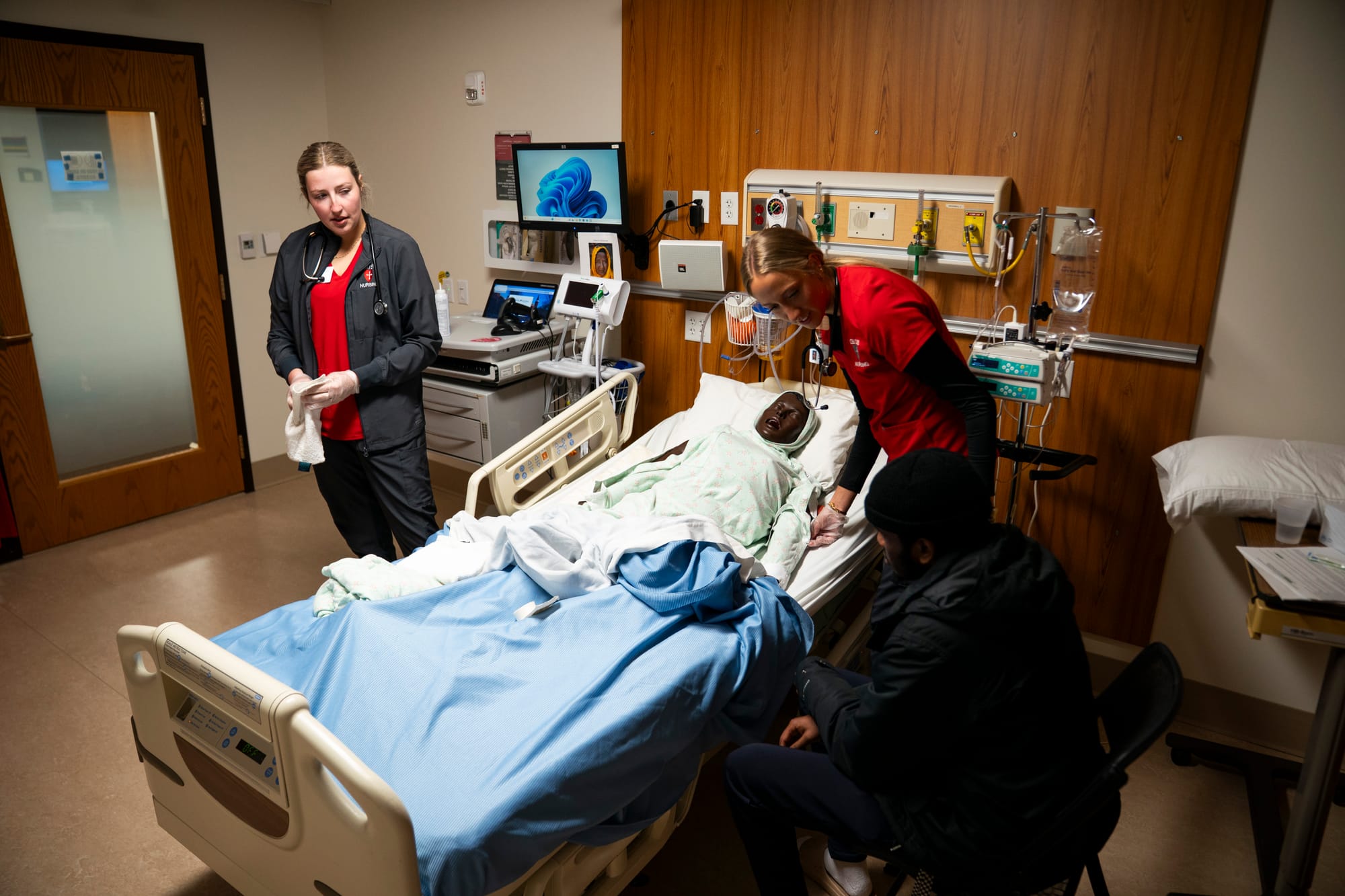 Two nursing students stand next to a hospital bed with a mannequin. They talk to a person acting as the patient's family during an end-of-life training exercise.
