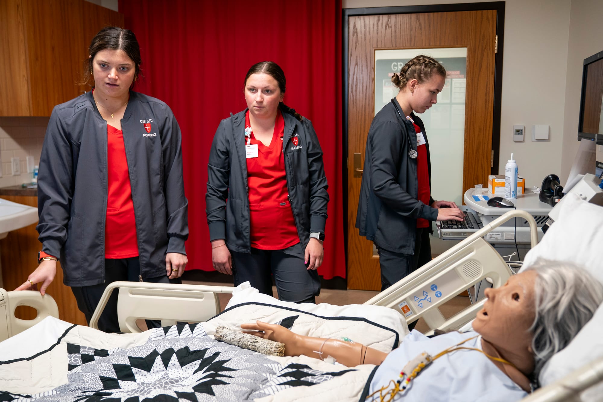 Three young women who are training to be nurses stand next to a patient's hospital bed. A mannequin lays in the bed during the training exercise.