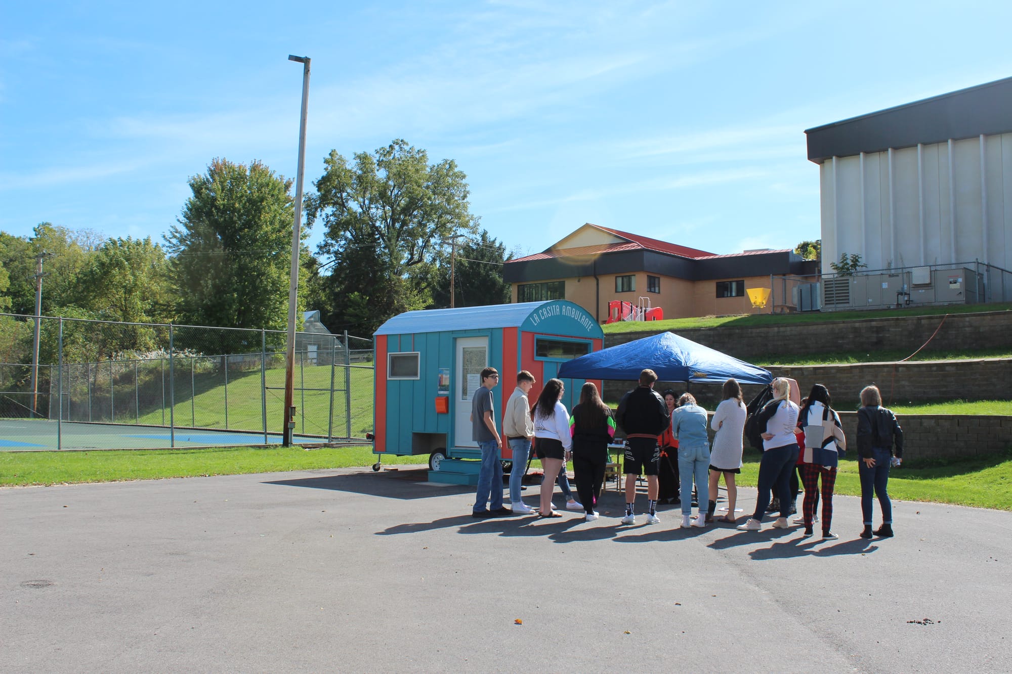 Ten people stand in line outside of "The Wandering House." They are on a paved surface with a basketball court behind them. 