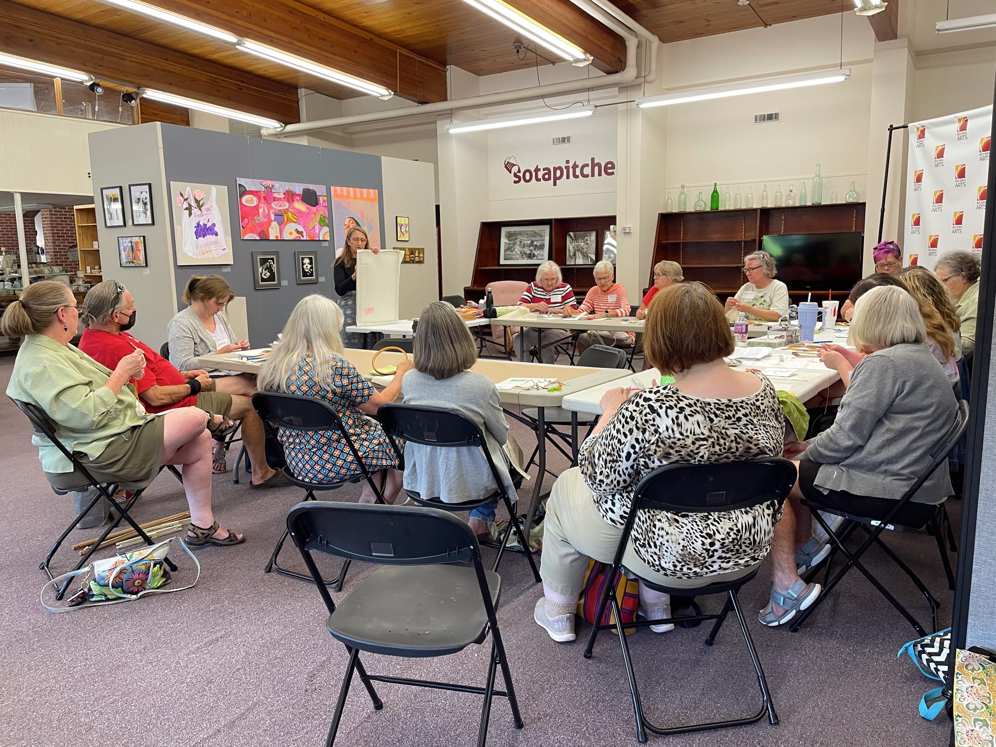 A total of 16 people — most of them women — sit in folding chairs doing embroidery work. Their heads are down as they concentrate on their work. 