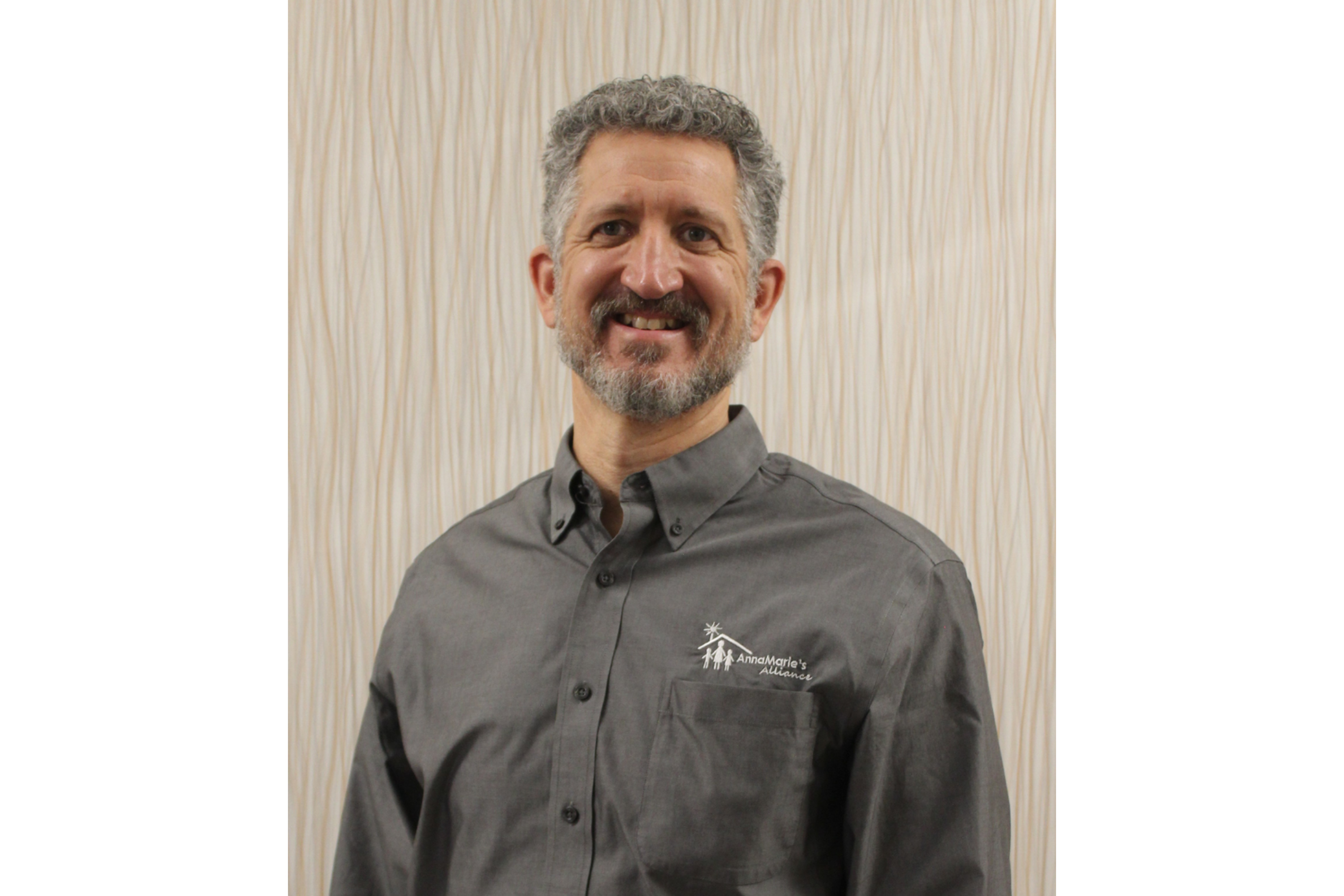 A man smiles for the camera. He wears a grey button-down shirt with the logo of Anna Marie's Alliance over the left breast pocket. 