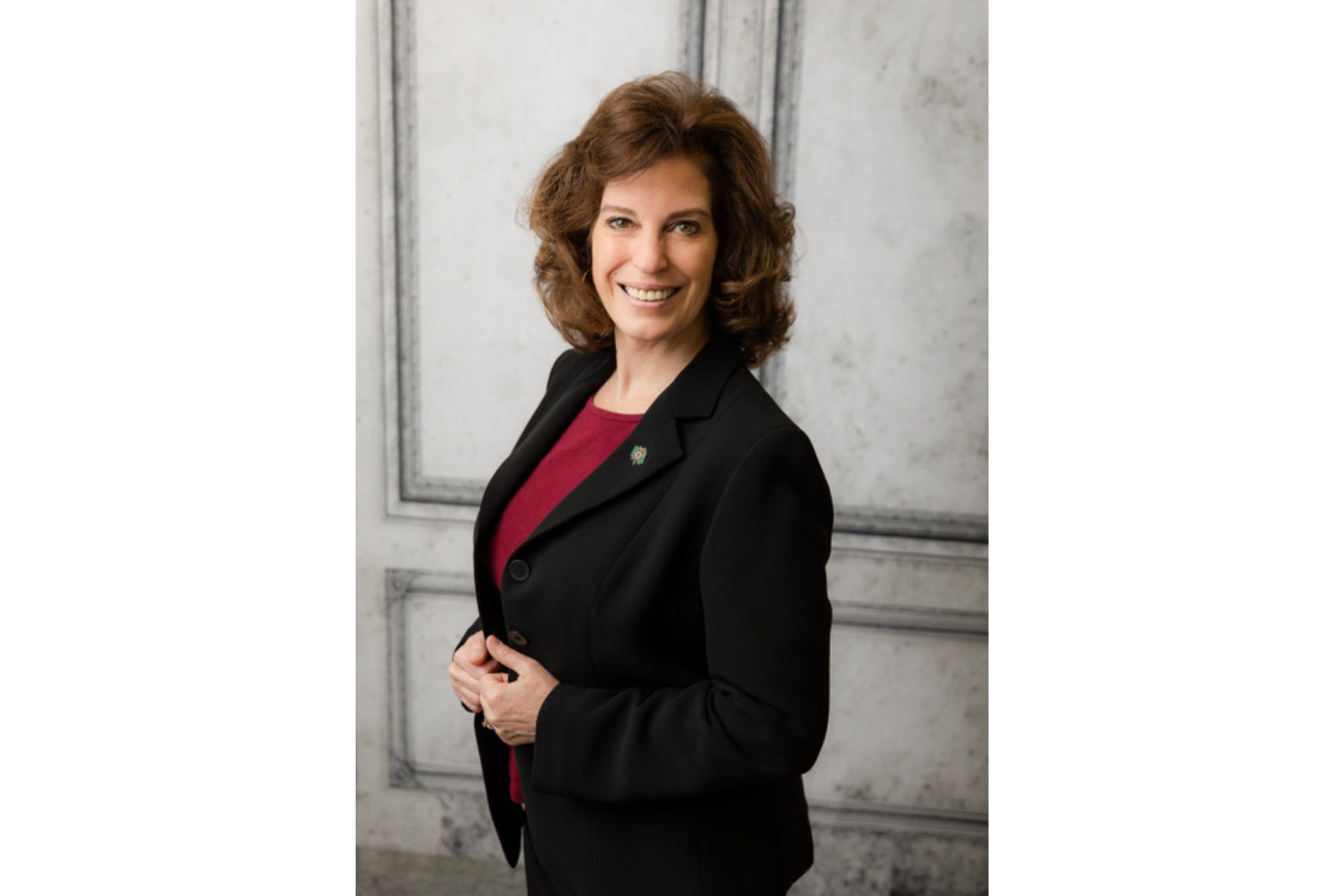 A woman smiles for the camera. She wears a red shirt under a black blazer. She has brown, curly hair. 