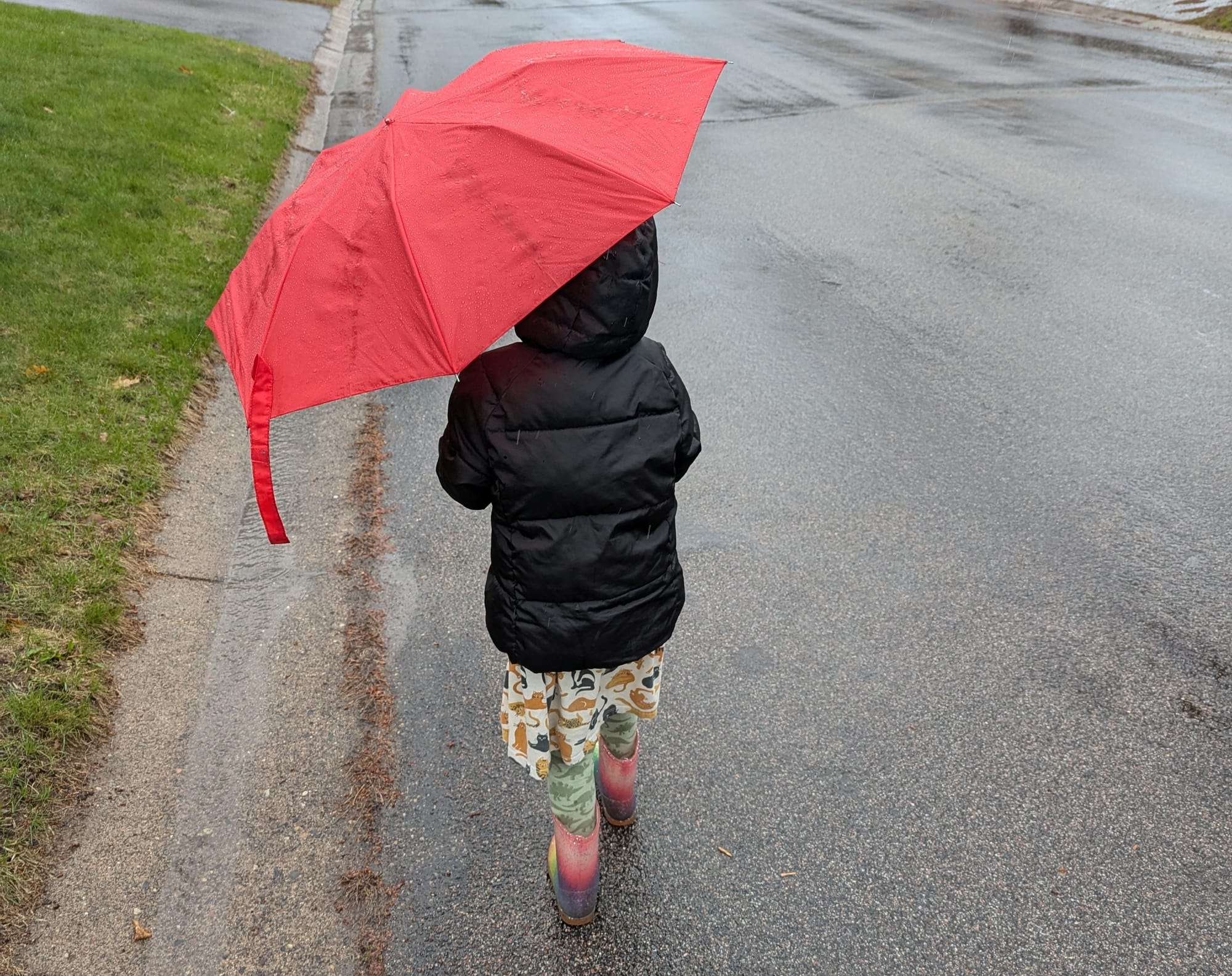 A child walks in the rain. They hold a red umbrella and wear a black raincoat with a skirt, leggings, and rainboots. 
