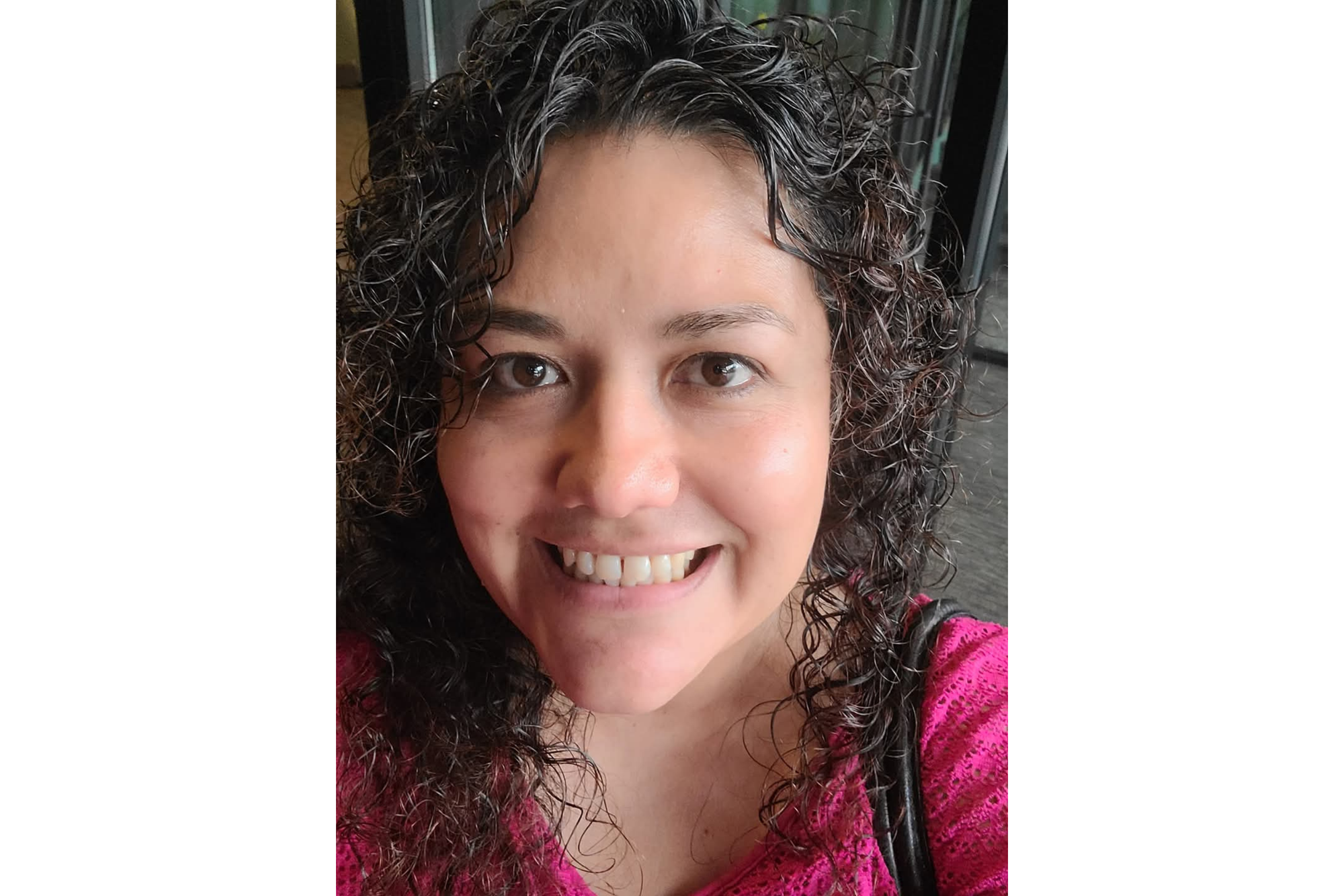 A selfie shows a woman smiling for the camera. She wears a pink shirt. Her wears her dark, curly hair down.