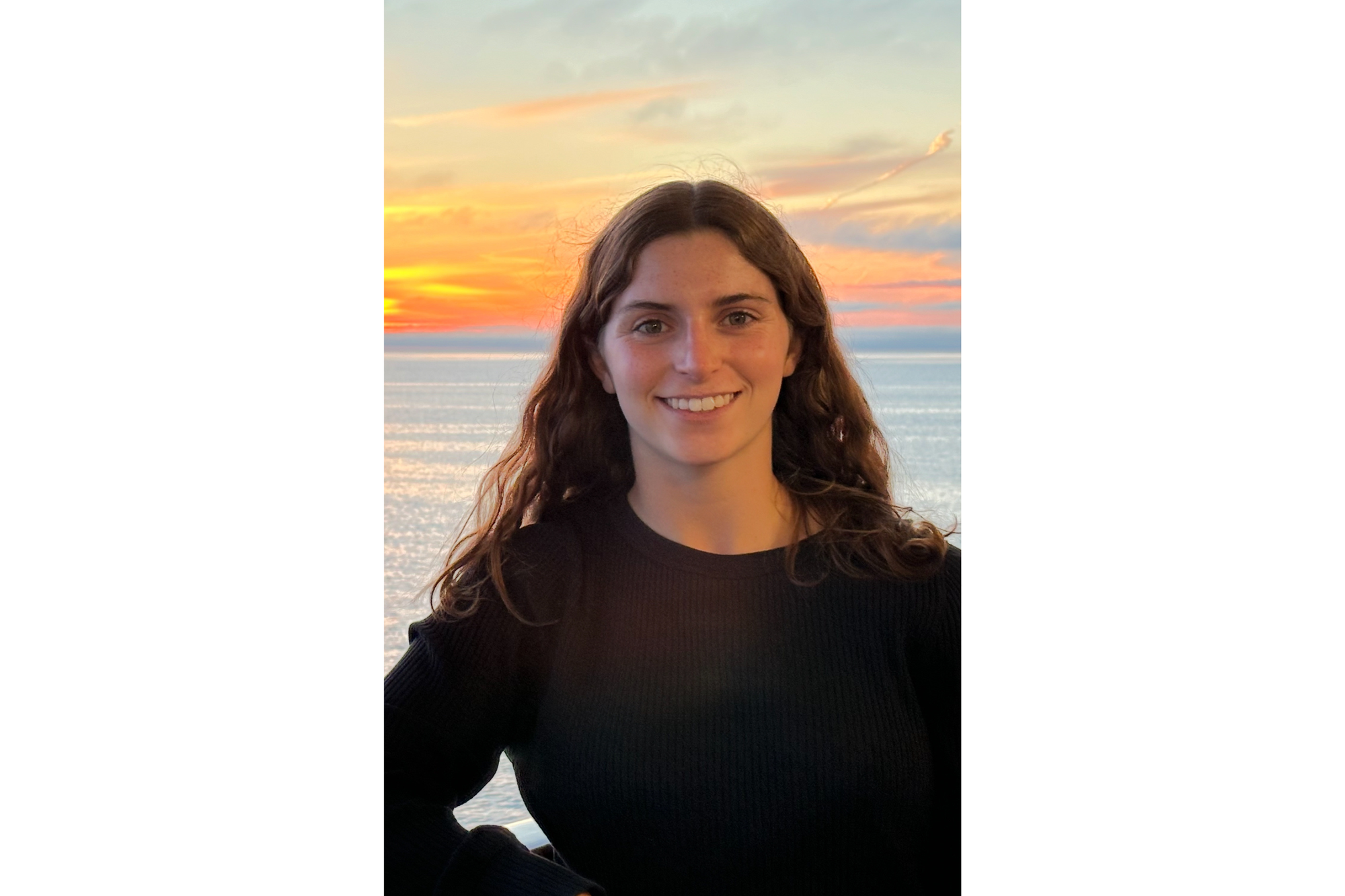 A young woman smiles for the camera. A blue lake is seen behind her along with an orange and yellow sunset. She wears a black long-sleeve shirt. Her brown, curly hair falls to her shoulders. 
