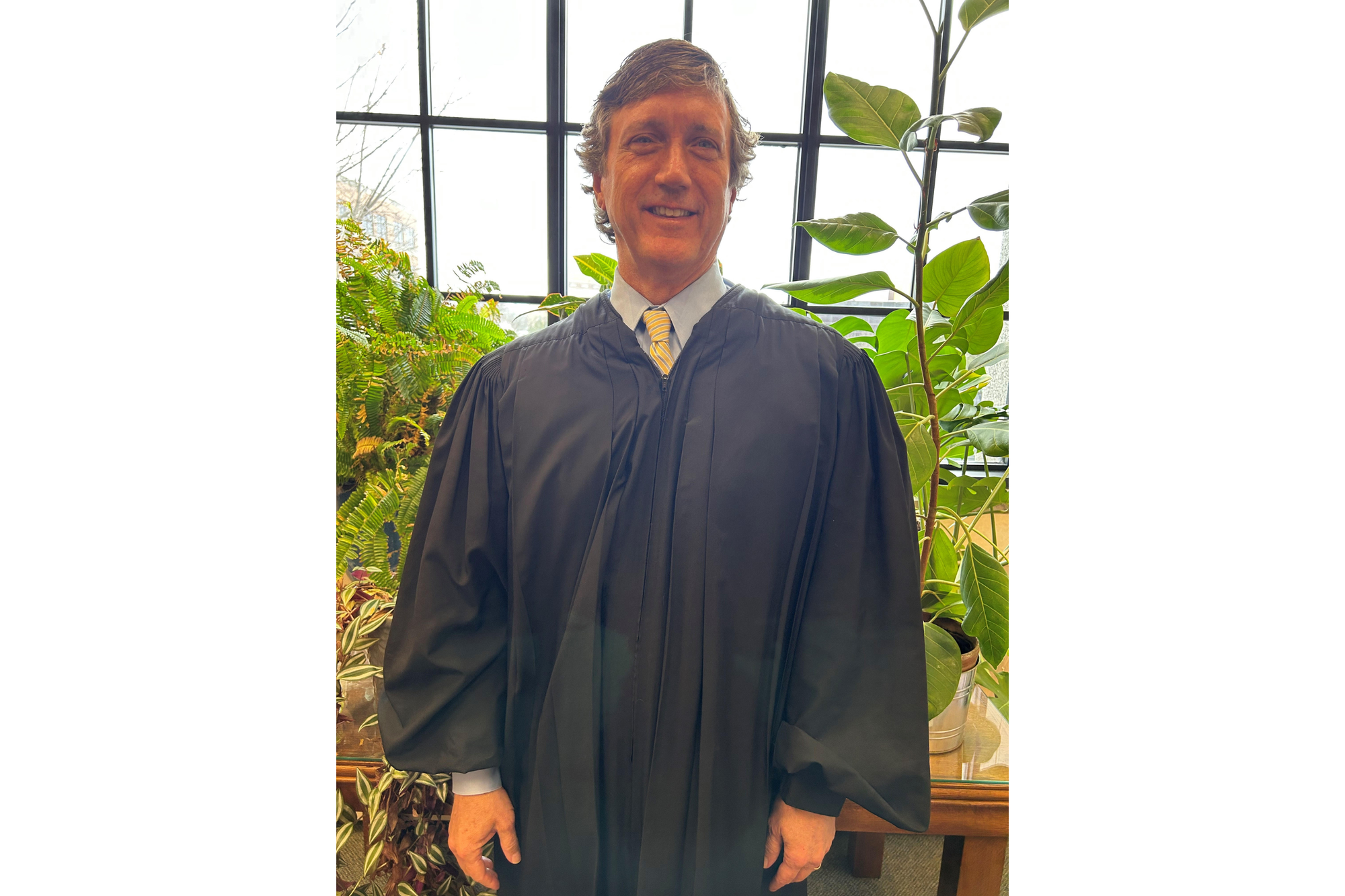 A man poses for a photo. He wears a blue button-down shirt with a yellow and blue striped tie. The shirt and top of the tie are visible under black judge's robes.