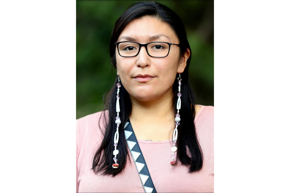 A young woman with dark hair and dark glasses poses for a photo. She wears a pink shirt and long earrings as long as her hair. 