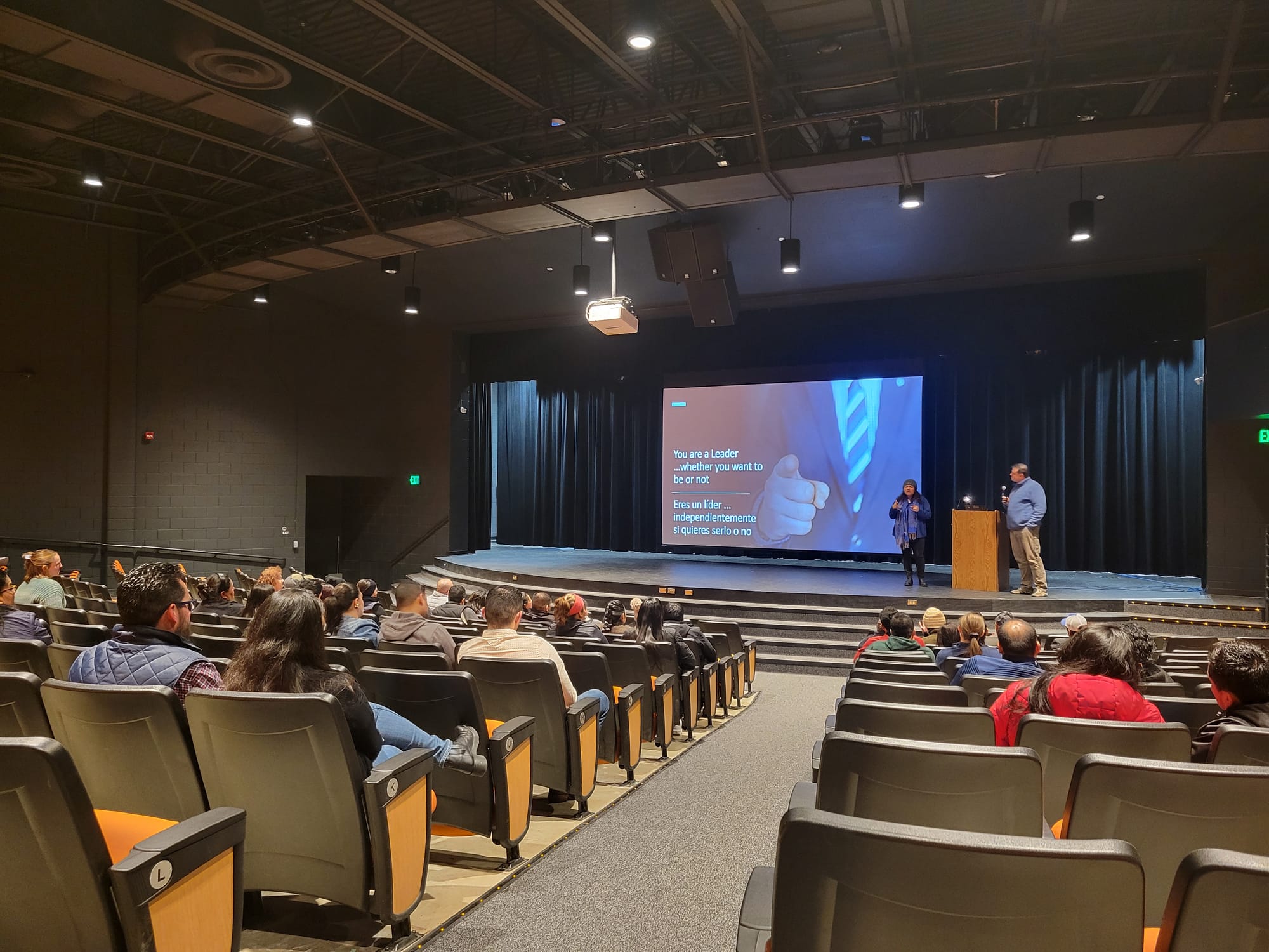 People listen to a presentation in an auditorium. A presentation is visible on a projection screen. Two people stand at a podium to the right of the screen.