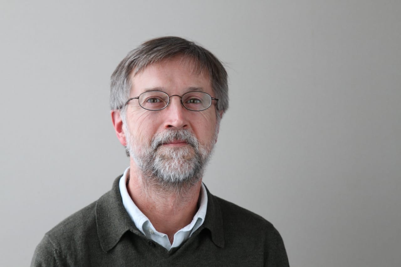 A man poses for a professional headshot. He wears a dark green sweater over a light blue collared shirt. He has a beard and wears glasses. 