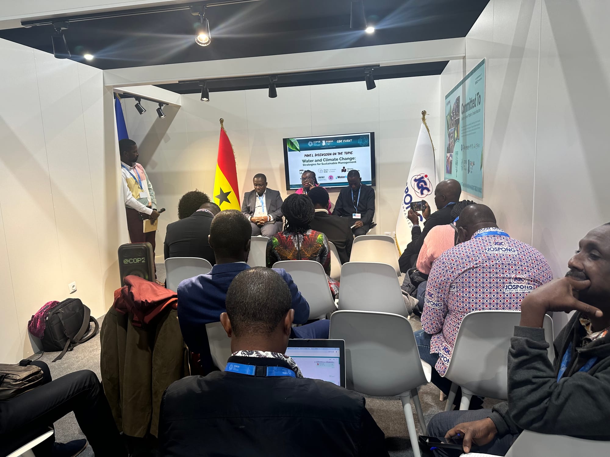 Nine audiences members are seen from behind during a panel discussion about water and climate change at COP29. Three panelists and a moderator are seen toward the back of the frame. 