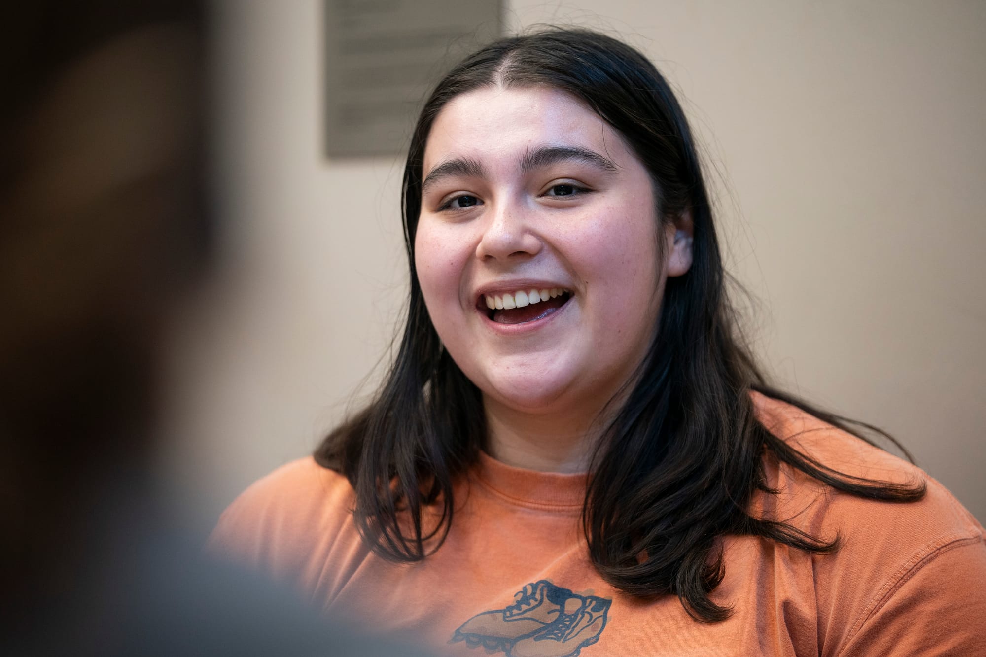 A young woman laughs during an event. She wears an orange T-shirt with a pair of boots on it. She has long brown hair. 