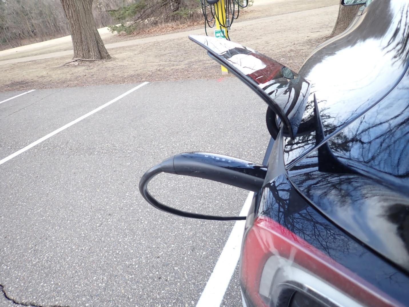 A black Tesla Model 3 charges at a nature center. Visible in the frame is the parking lot and a few trees in the background. 