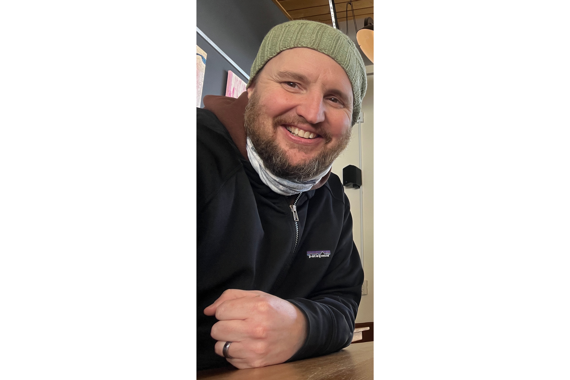 A man smiles for a selfie. He wears a black sweatshirt and a green stocking cap. He has a beard. 