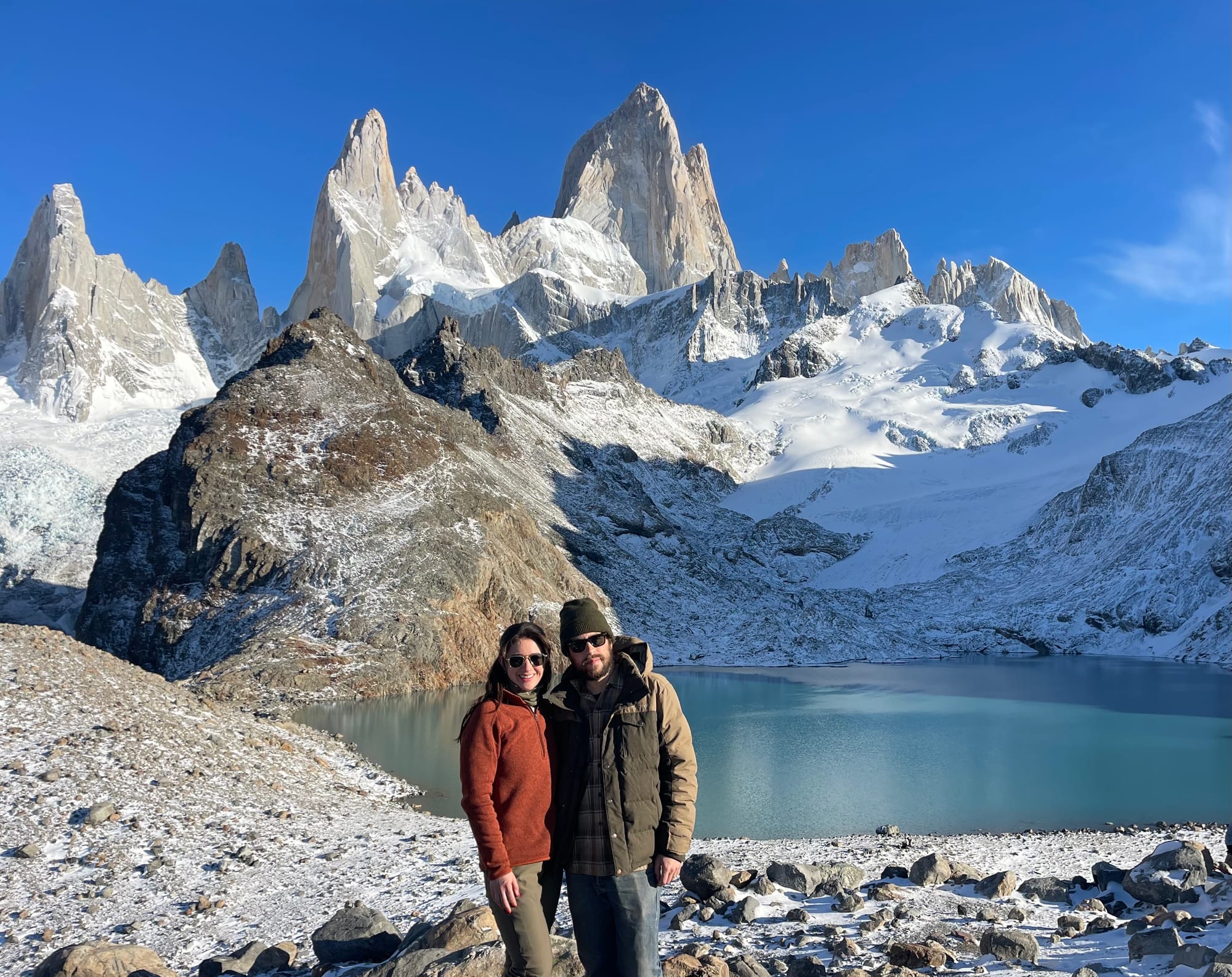 A couple pose for a photo in the snowy mountains of Patagonia. Behind them is a blue lake and snow-covered peaks. She wears a red sweatshirt with brown pants and sunglasses. He wears a brown coat with jeans, sunglasses and a stocking cap. 