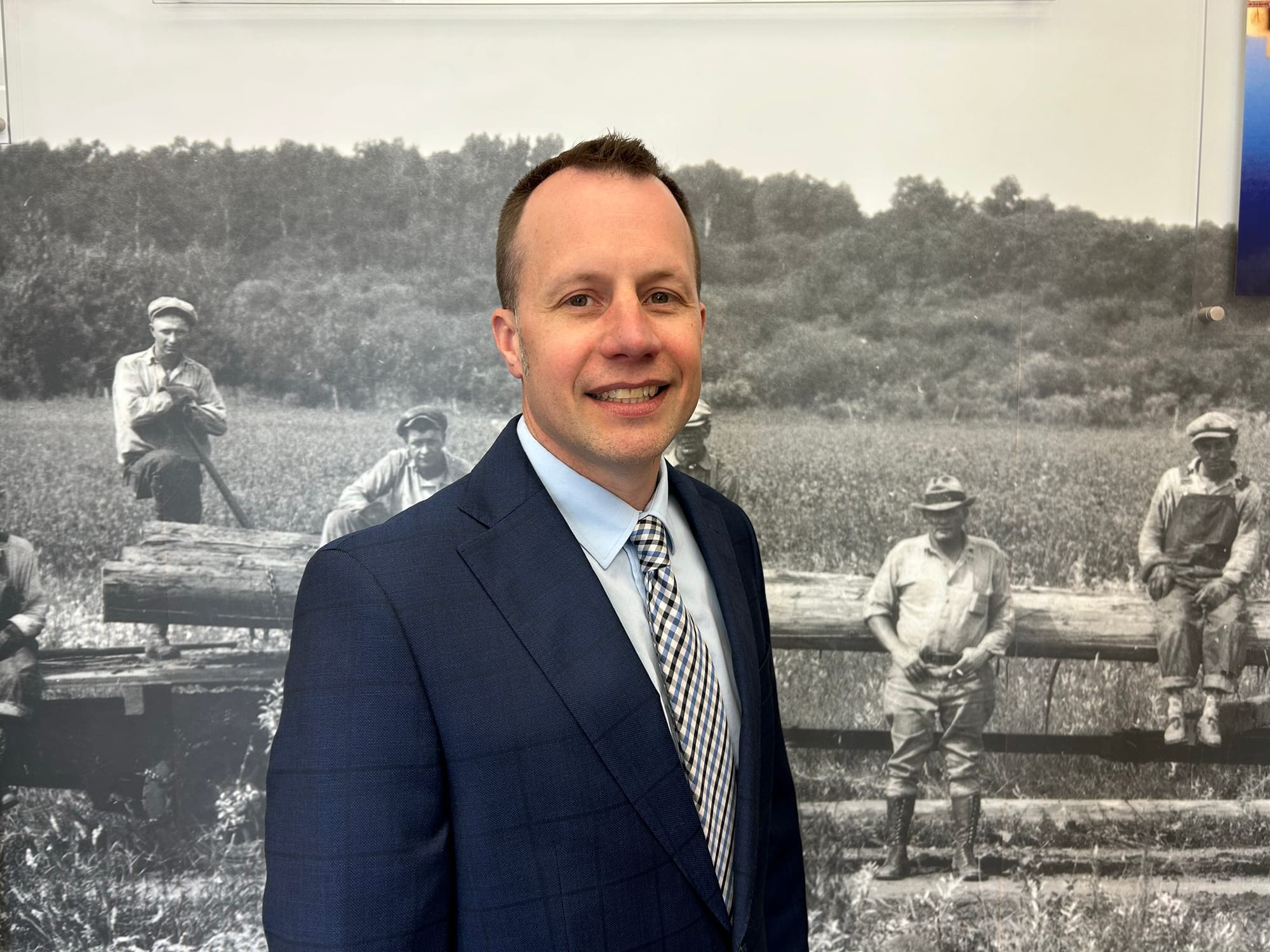 A man smiles for a photo. He wears a blue suit jacket over a light blue shirt with a striped tie. He stands in front of an historical, black and white photo. 