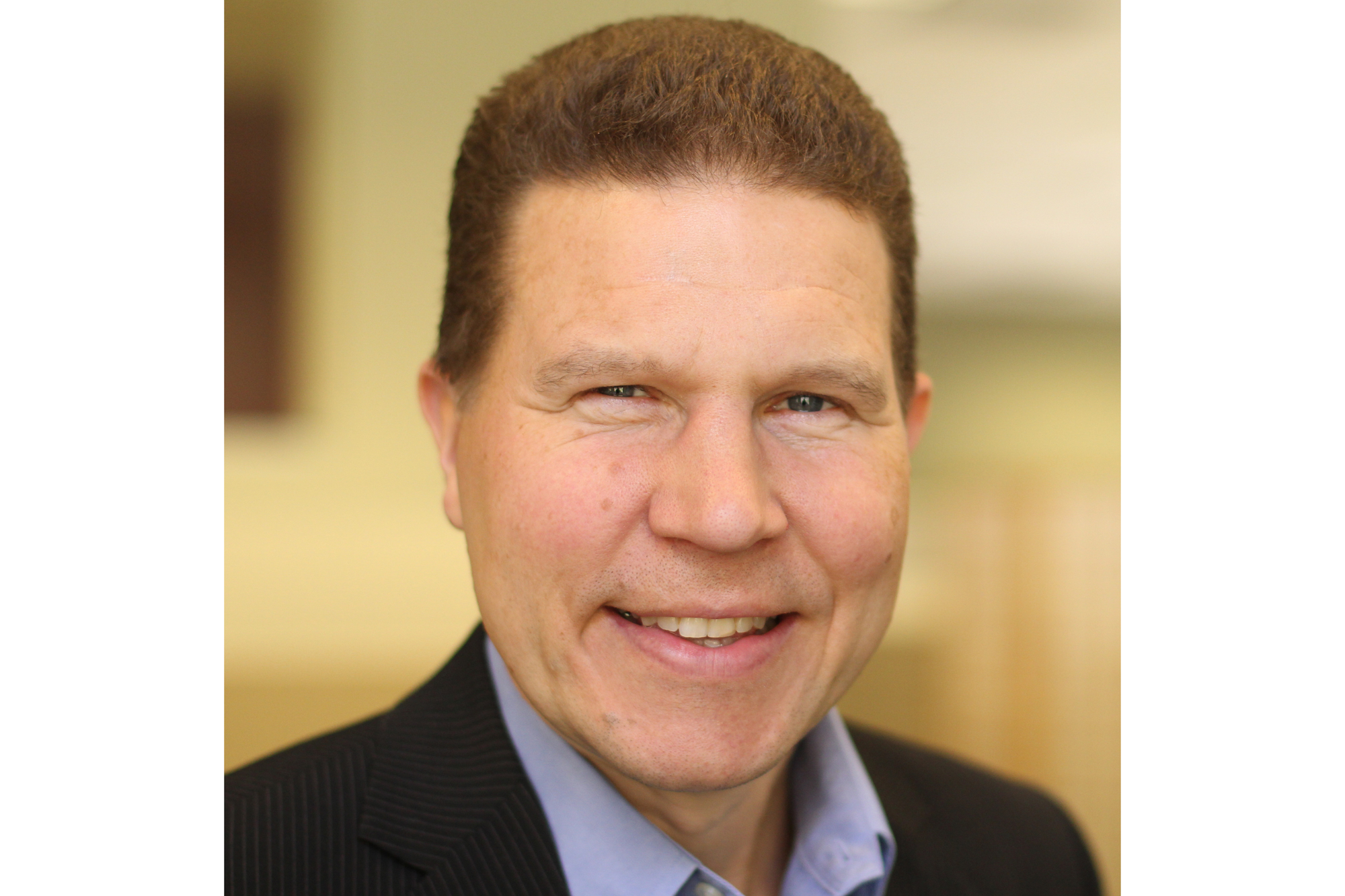 A man smiles for a headshot. His head and shoulders are visible. He wears a dark suit jacket with a bluish-purple shirt. He has short, brown hair. 