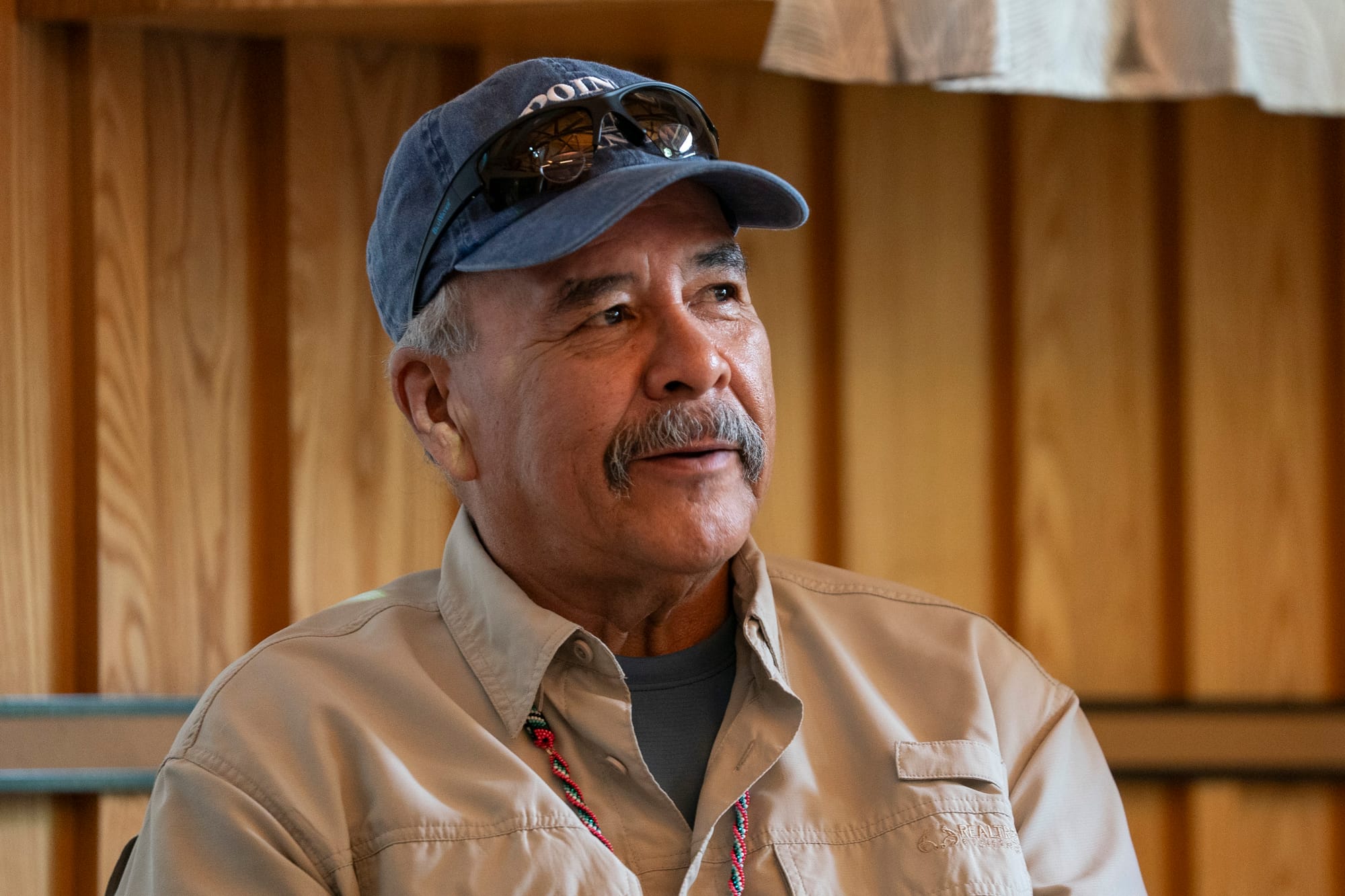 A Native American man smiles during an event. He wears a blue baseball cap with sunglasses on the brim. Gray hair peeks out underneath the hat near his ears. His mustache is dark with flecks of gray. He wears a tan button-down shirt over a blue T-shirt. He wears a beaded necklace. 
