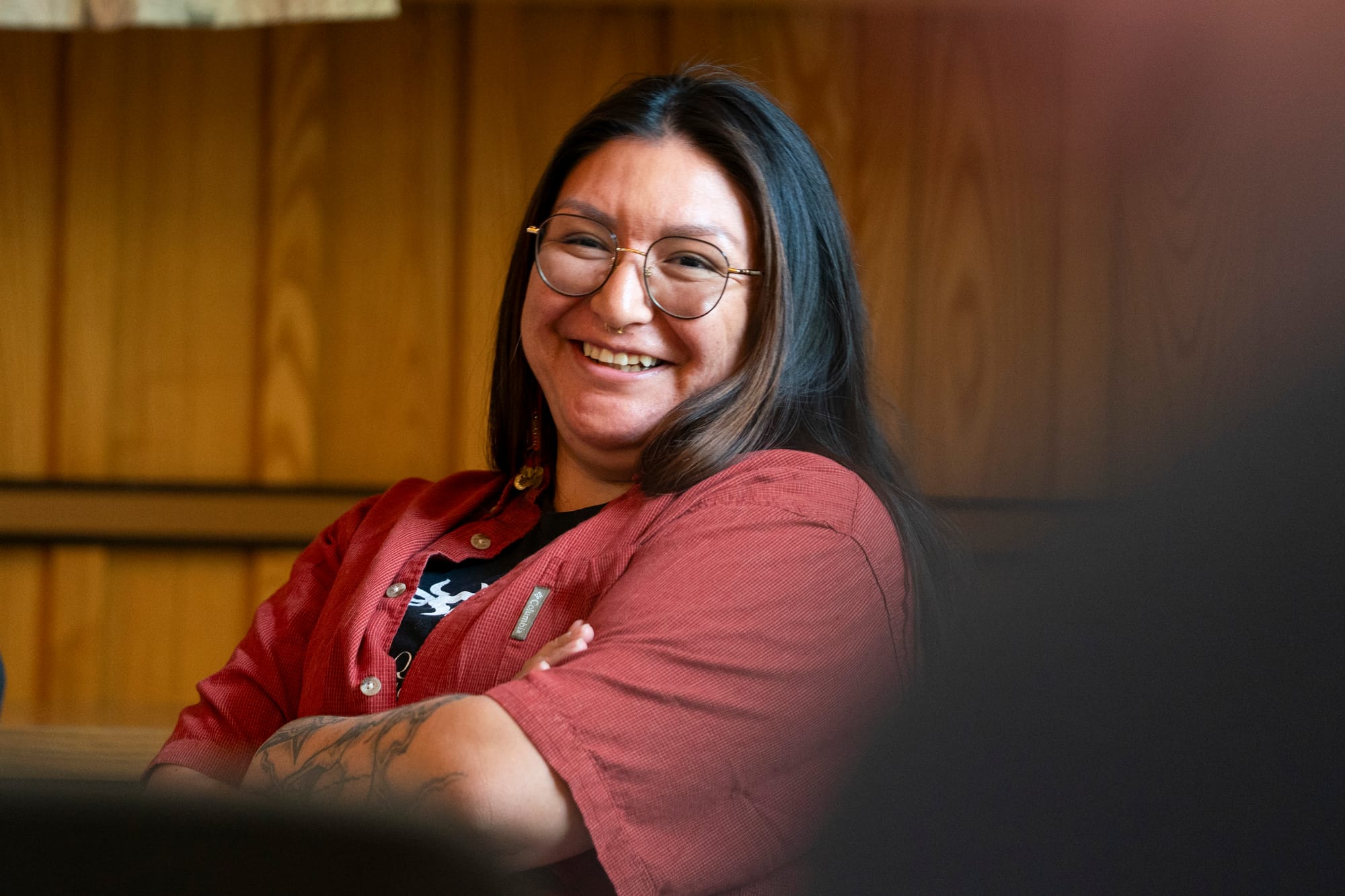 A Native American woman smiles during an event. She has long, dark hair that falls past her shoulders. She wears glasses and has a nose ring. She wears a red, short-sleeve button-down shirt over a black T-shirt. Tattoos are visible on one of her arms. 