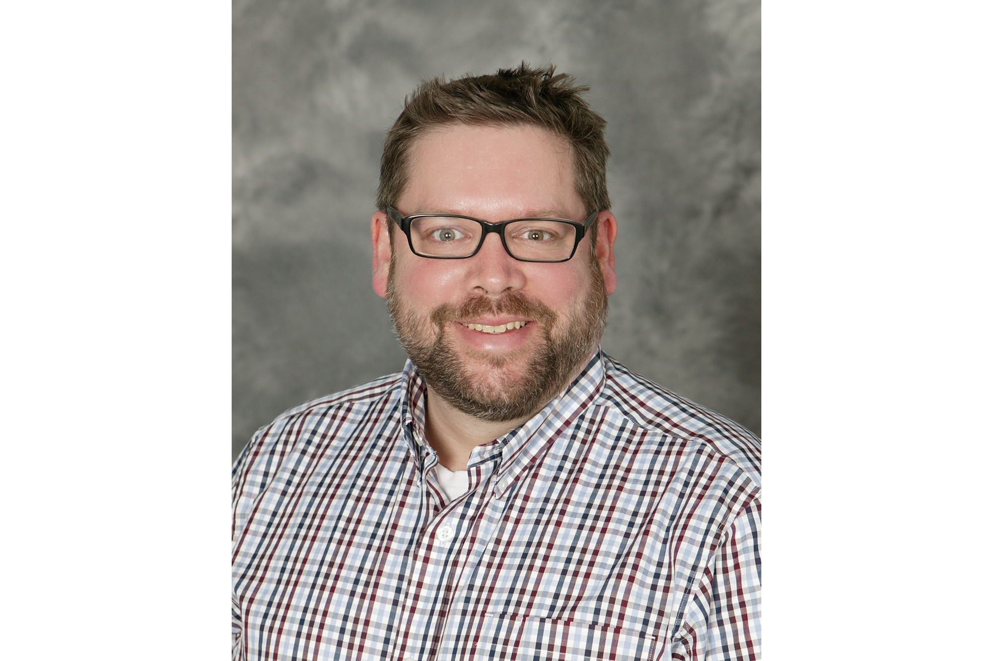 A white man poses for a professional headshot. He has brown hair, a brown beard, and wears black glasses. He wears a striped button-down shirt. Behind him is a gray backdrop. 