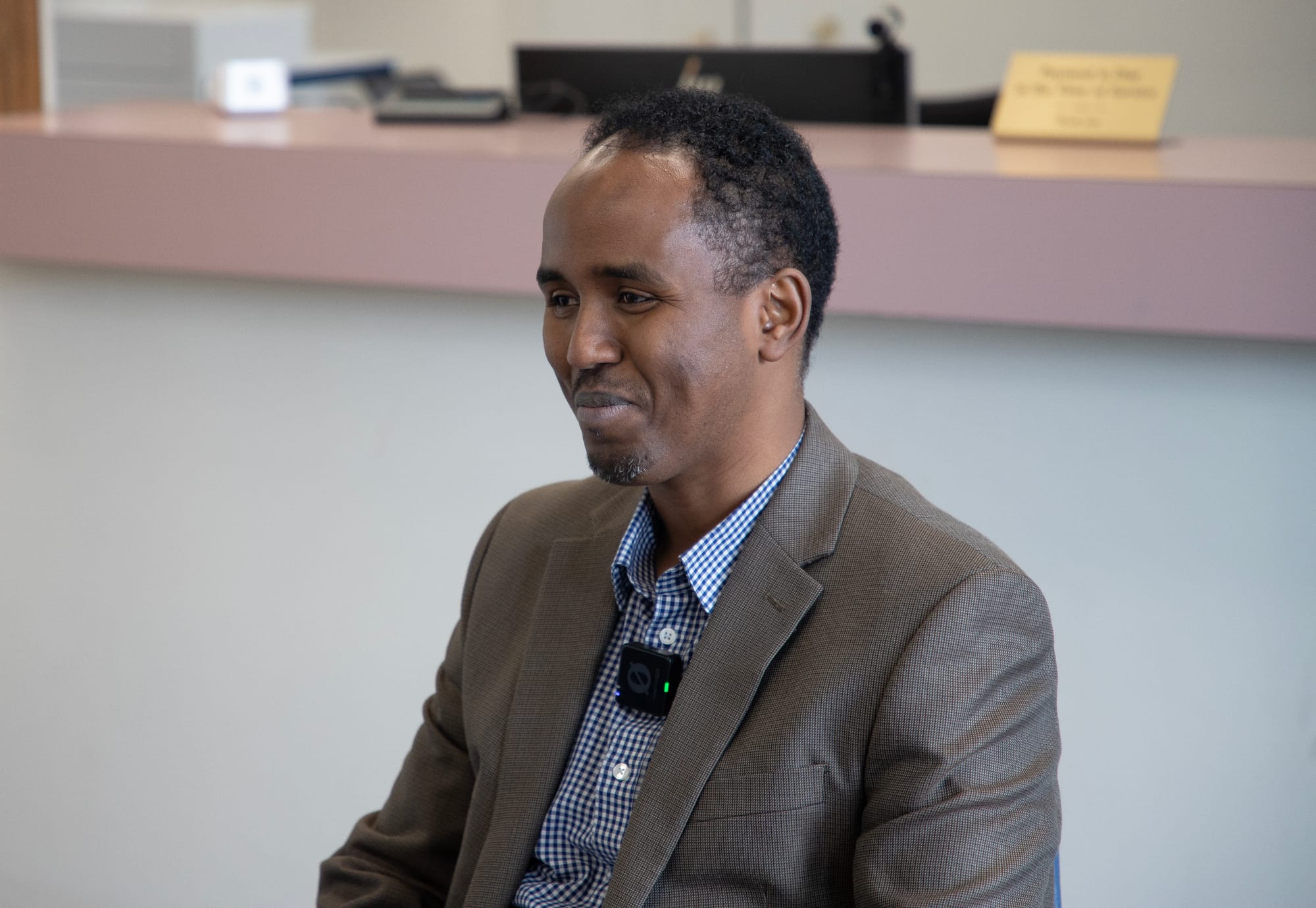 Nin dhoola cadaynaya xili aan waraysigga qaadaynay. Dhanka bidixda ee sawirka. Wuxuu xiran yahay shaati buluug ah oo jacket midab cawlan. Wuxuu leeyahay timo gaaban oo madow. 