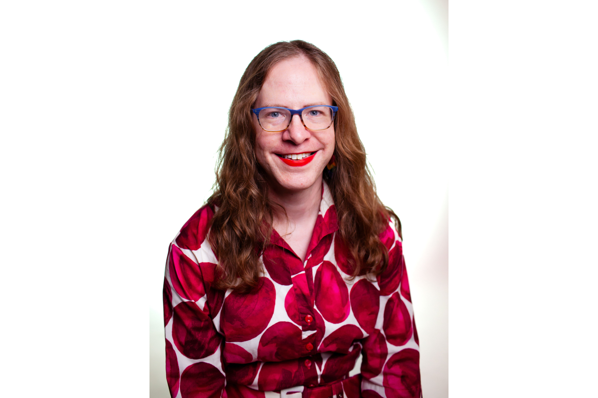 A person poses for a professional portrait. She wears a white blouse with bright red polka dots. She wears her reddish-brown hair down - it falls past her shoulders. She also has blue glasses on. She smiles for the camera. 