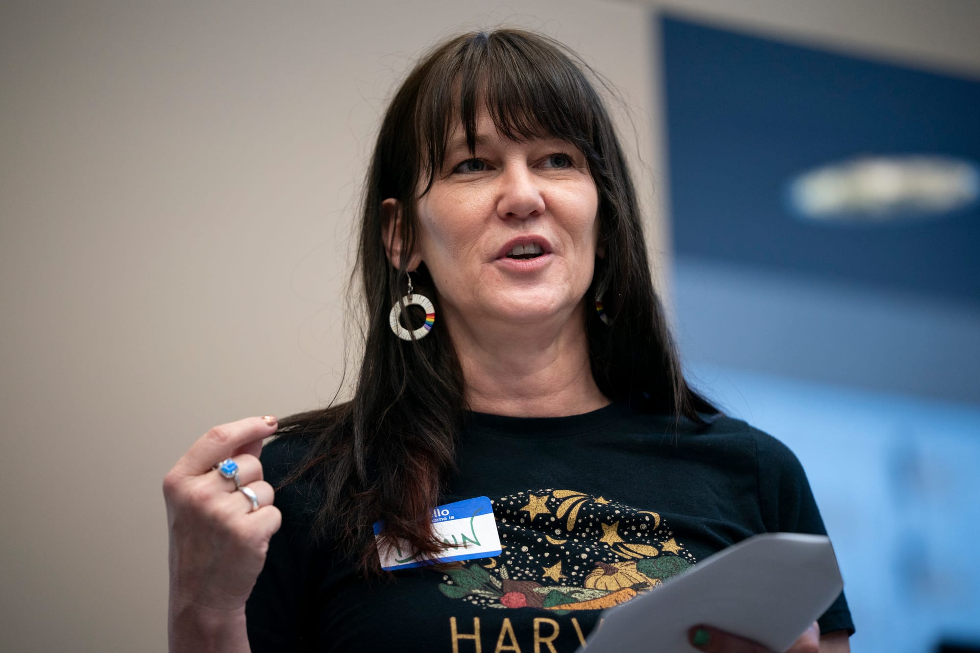 A woman speaks to a crowd. She has long dark hair and bangs. She wears a black t-shirt with a white and blue name tag. Her white hoop earrings have the rainbow pride colors visible. 