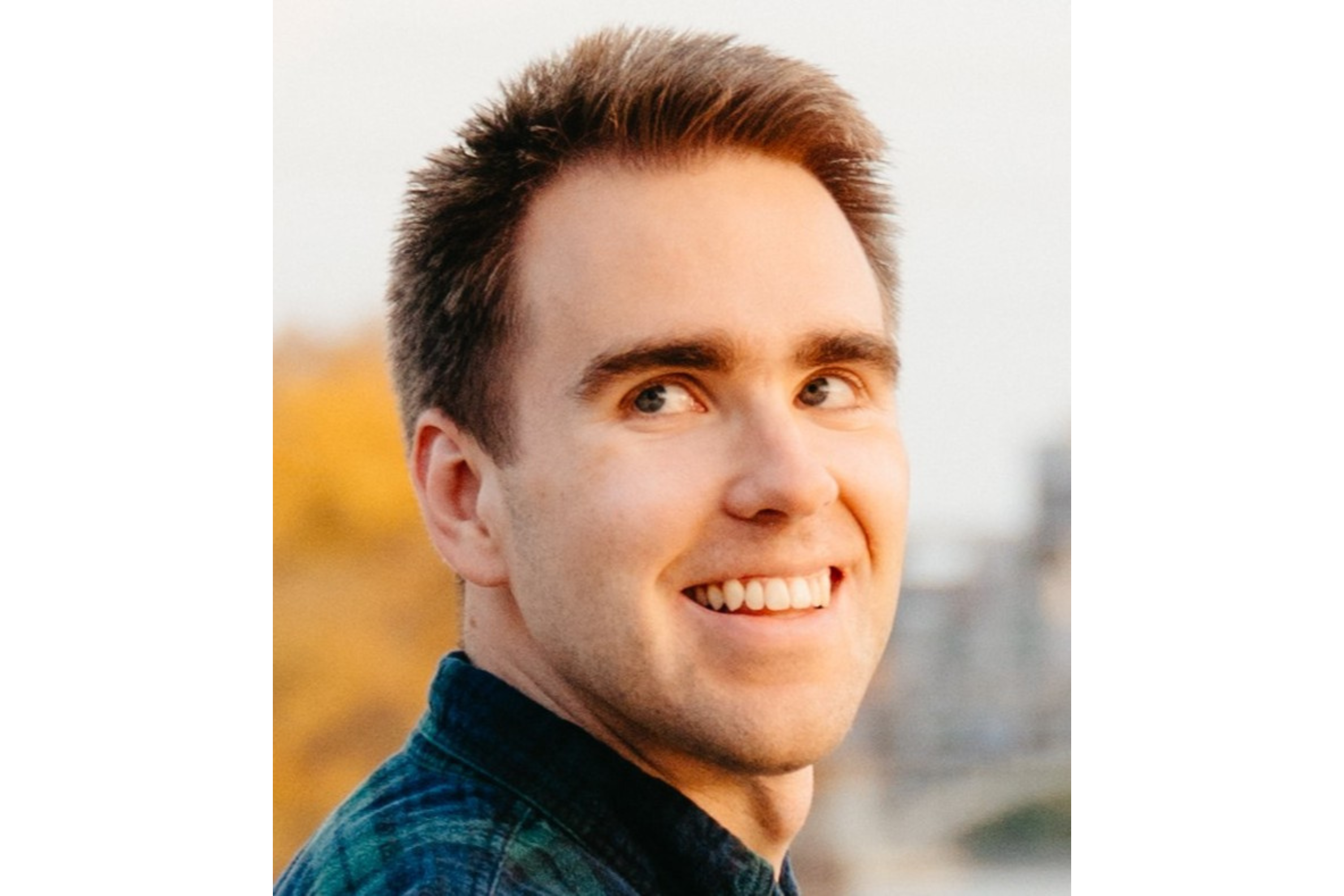 A man smiles as he looks to the upper left corner of the frame. He wears a green and blue plaid flannel shirt. He has short brown hair. 