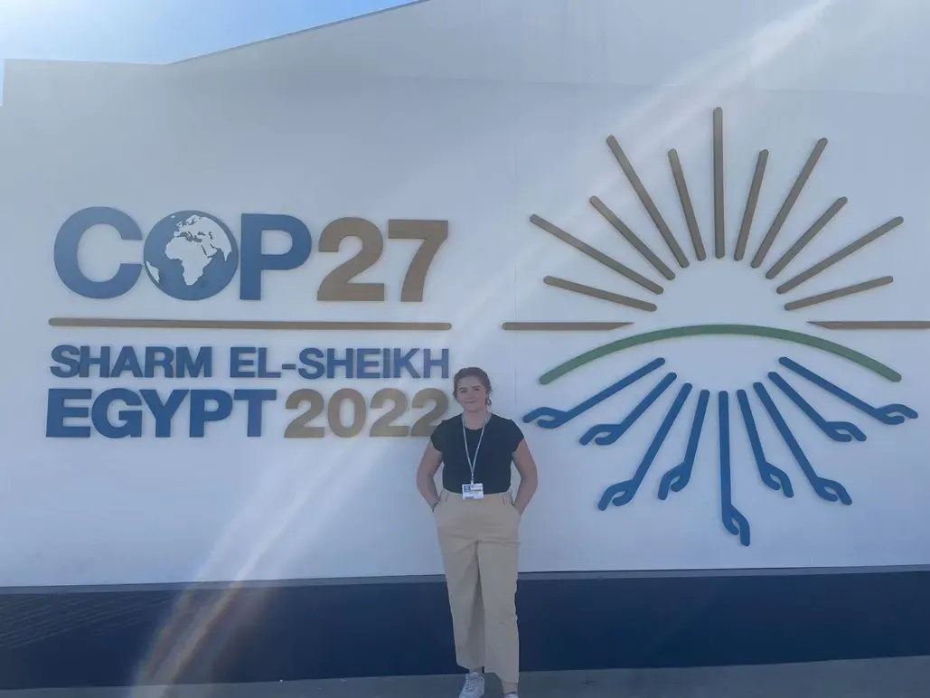 Photo of college student Claire Boettcher in front of a sign reading: COP27, Sharm el-Sheikh, Egypt 2022