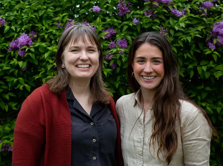 Two women with dark hair smile as they pose for a photo in front of lilac bushes. 