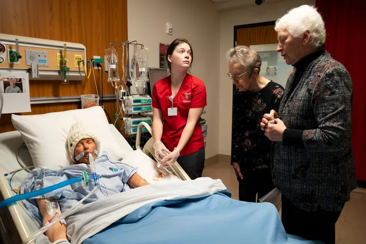 A nursing student administers medication to a mannequin patient while two people say their goodbyes during a training exercise. 