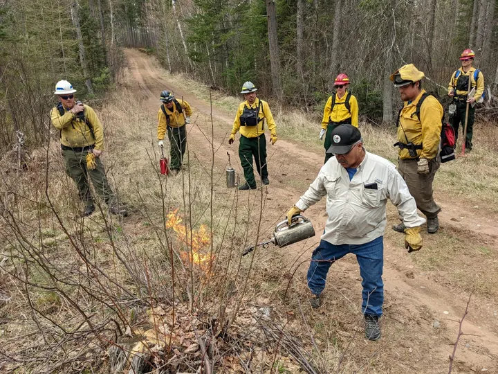 A man lights a fire for a prescribed burn while firefighters watch. 
