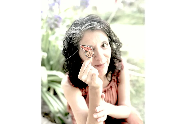 Una mujer sostiene un ala de mariposa monarca sobre uno de sus ojos mientras mira a la cámara. Lleva un vestido naranja quemado. Tiene el pelo oscuro y rizado. 