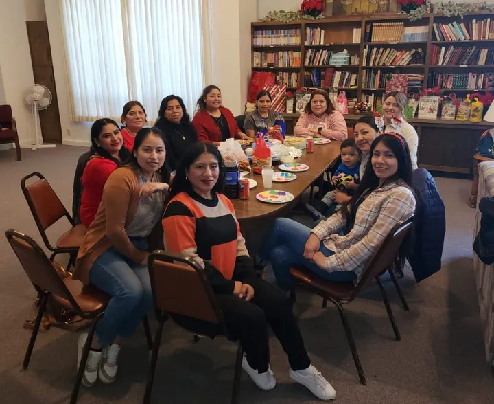 Eleven Latina women sit around a table and face the camera. Many are smiling. They are gathered for a support group. 