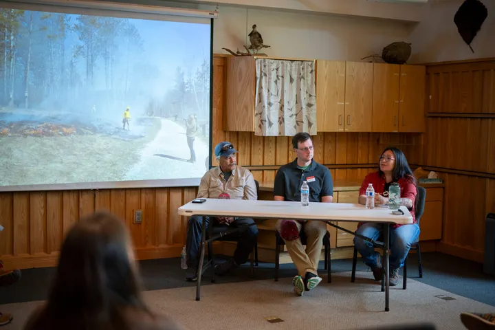 Three people sit at a white folding table. The woman on the right is speaking. The two men next to her listen. In the background is a projection screen with an image of a controlled burn. 