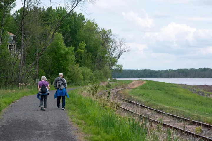 Two people walk along a paved trail. Railroad tracks are seen to their right, with the river bank even further to the right of the frame. 