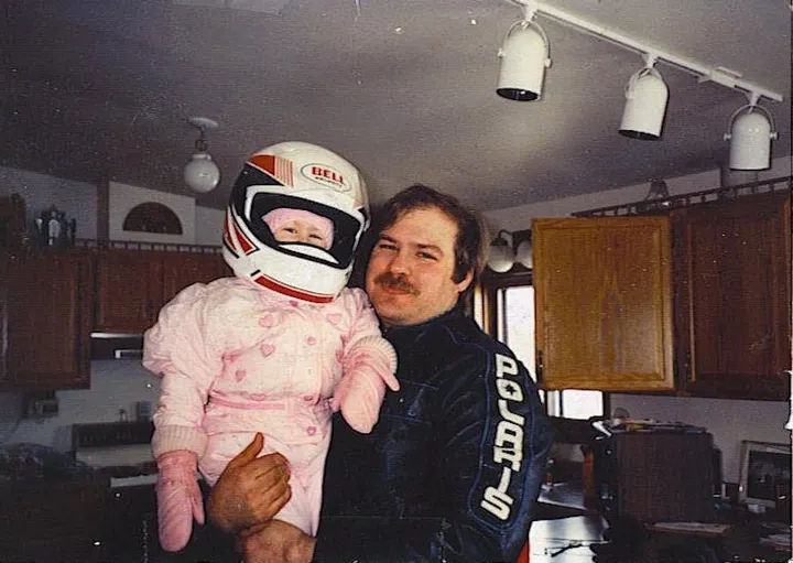 A man in his 30s with brown hair and a brown mustache holds a little girl. She's bundled up in a pink snowsuit with pink mittens. She wears a snowmobile helmet. 