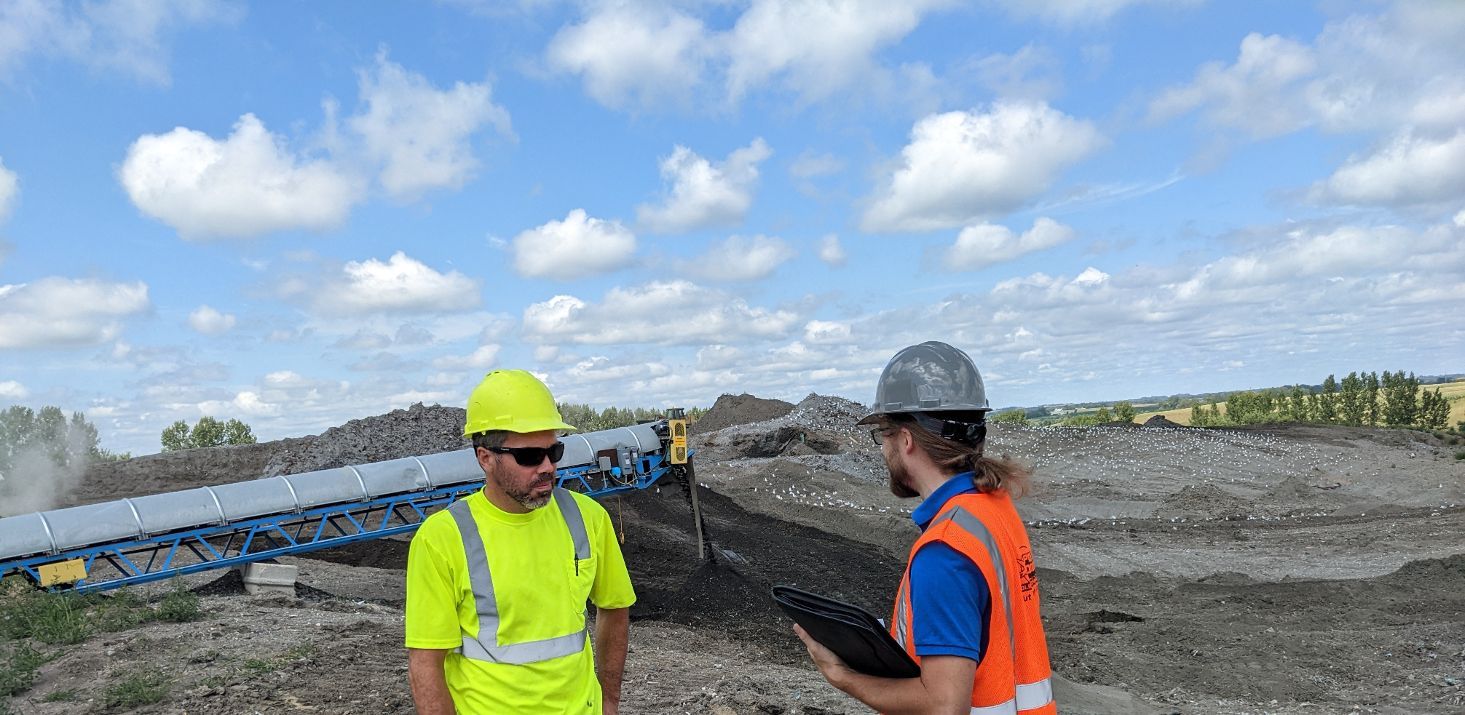 Ash is returned via conveyor belt to the landfill after passing through Pope/Douglas Solid Waste Management's Metal Recovery Facility in Hoffman, Minn., on Tuesday, Aug. 16, 2022, as Facility Manager Nick Trinka (left) talks with visitors during a site visit. The white dots in background are seagulls picking through the ash landfill. (Nora Hertel for Project Optimist)