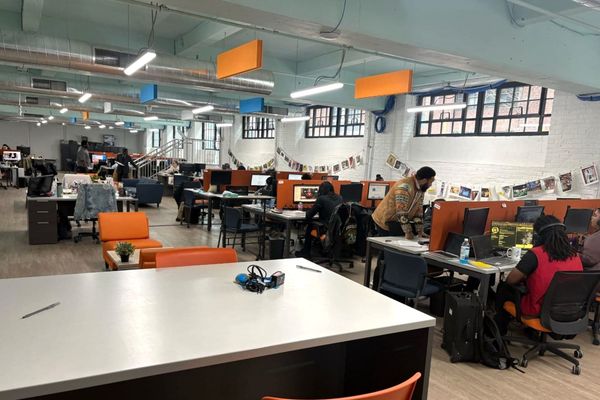 A staff member stands beside seated trainees at computer workstations in a bright room with white brick walls.