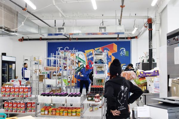 Inside Greater Goods in Kensington. Shoppers and volunteers move among shelves of juice, peanut butter, sauces and fresh produce. A blue mural for The Sunday LOVE Project covers the wall.