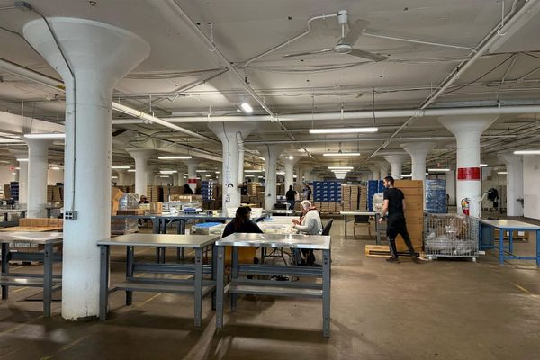 A wide view of the Baker Industries production floor in Kensington, with workers at stations between tall white columns and stacks of cardboard boxes in the background.