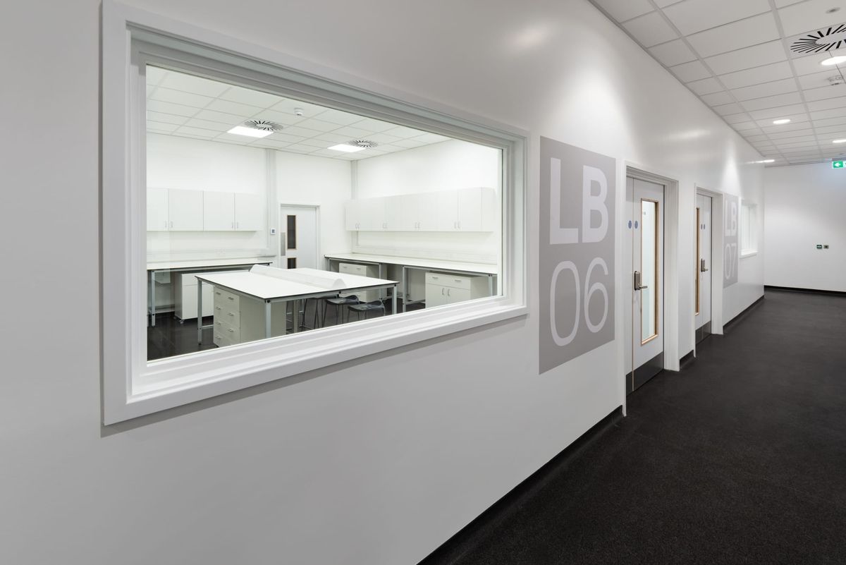 View through a large internal window into a clean, white laboratory room labeled LB06 at the Science Creates Old Market incubator, featuring lab benches, storage cabinets, and stools.