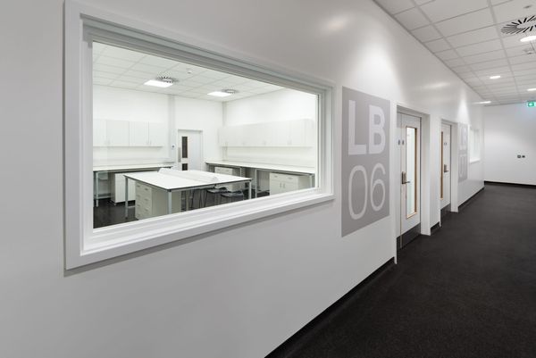 View through a large internal window into a clean, white laboratory room labeled LB06 at the Science Creates Old Market incubator, featuring lab benches, storage cabinets, and stools.